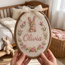 A person holds an oval embroidered hanging piece in a wooden hoop in a nursery setting. The embroidery features a pink bunny holding a heart, a butterfly, floral details, and the name 'Olivia' in pink script, with a cream lace bow at the top.