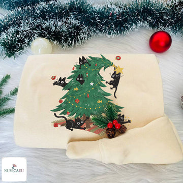 A folded cream-colored sweatshirt featuring a festive Christmas tree design with black cats climbing and hanging around the tree, styled on a white faux fur surface with tinsel and ornaments.
