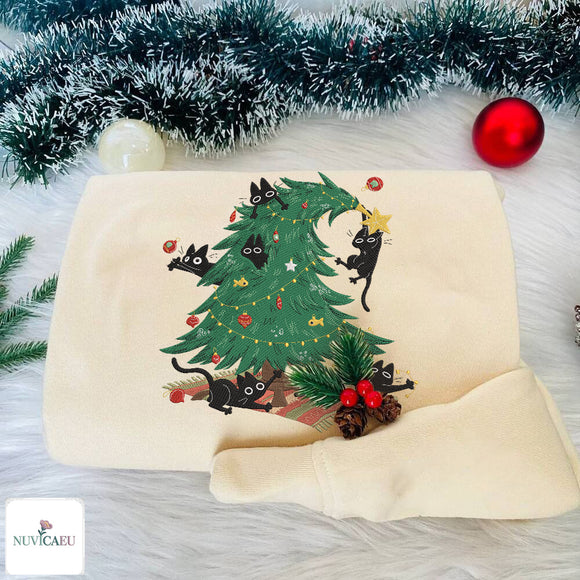 A folded cream-colored sweatshirt featuring a festive Christmas tree design with black cats climbing and hanging around the tree, styled on a white faux fur surface with tinsel and ornaments.