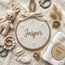 A styled baby nursery flat lay featuring a personalized wooden hoop ornament embroidered with the name 'Jasper' and white daisy flowers, surrounded by a cream crochet bunny, wooden and silicone pacifiers, a beaded ring teether, a plush sock, and a swaddle blanket with a matching wooden milestone insert.