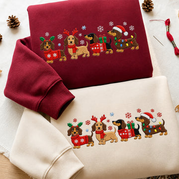 Two folded sweatshirts with matching Christmas dachshund embroidery, one in burgundy and one in cream, styled on a light fabric background with pinecones and thread.