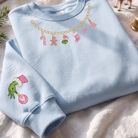 A light blue crewneck sweatshirt laid flat on a white fabric surface. The sweatshirt features a gold chain graphic across the chest with Christmas-themed charms in pink, green, and brown, and an embroidered Grinch motif on the sleeve.