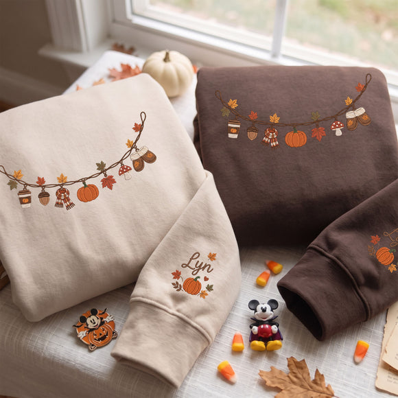 Three folded sweatshirts with autumn-themed embroidery, including pumpkins, acorns, leaves, and a cursive name, styled on a table with candy corn, a small pumpkin, and a Mickey Mouse charm.