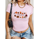 A woman wearing a light pink short-sleeve t-shirt with a cozy autumn-themed graphic print that reads 'Cozy season' surrounded by pumpkins, acorns, a campfire, and a beanie. She is also wearing denim shorts and a black shoulder bag.