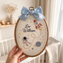 A hand holds an oval embroidered canvas in a wooden hoop, featuring a light blue satin bow at the top. The embroidery shows a space theme with an astronaut, a rocket ship, stars, and the personalized name 'Leo William' in dark blue script.