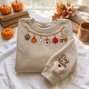A folded beige crewneck sweatshirt on a white fabric surface, decorated with a pumpkin-themed chain graphic and 'Dog Mom' text on the sleeve. The scene is styled with autumn props including pumpkins, books, a mug, and a scarf.