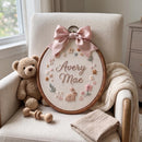 A personalized embroidered wooden hoop wall hanging with a pink satin bow, displayed on a cream upholstered armchair next to a teddy bear and a folded blanket.