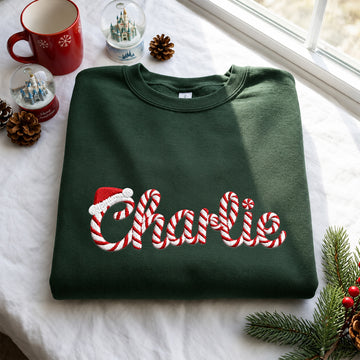 A dark green crewneck sweatshirt with 'Charlie' embroidered in red and white candy cane script and a Santa hat, displayed on a white blanket with Christmas decorations.