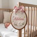 A personalized baby nursery wall hanging in an oval wooden hoop, featuring a pink satin bow, embroidered name 'Adeline', a white bunny, flowers, and butterflies. The hoop is hung on a light wood crib with a sleeping newborn baby in a white swaddle blanket in the softly blurred background.