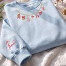 A light blue crewneck sweatshirt laid flat on a white fabric surface, decorated with a gold chain graphic featuring Christmas motifs and pink text reading 'Amee' on the sleeve.