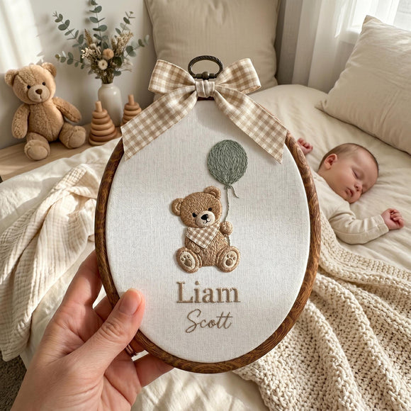 A hand holds an oval embroidery hoop featuring a personalized embroidered teddy bear holding a balloon with the names 'Liam Scott' stitched below. The hoop is decorated with a beige and white gingham ribbon bow at the top.