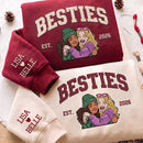 Three folded sweatshirts arranged on a light fabric background: a burgundy sweatshirt with cream 'BESTIES EST. 2026' text and a cartoon hug graphic, a cream sweatshirt with burgundy 'BESTIES EST.