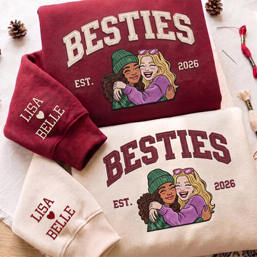Three folded sweatshirts arranged on a light fabric background: a burgundy sweatshirt with cream 'BESTIES EST. 2026' text and a cartoon hug graphic, a cream sweatshirt with burgundy 'BESTIES EST.