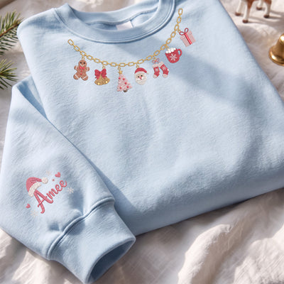 A light blue crewneck sweatshirt laid flat on a white fabric surface, decorated with a gold chain graphic featuring Christmas motifs and pink text reading 'Amee' on the sleeve.