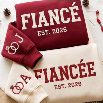 Two embroidered sweatshirts, one burgundy and one cream, laid flat on a light surface with decorative pinecones and thread. The burgundy sweatshirt reads 'FIANCÉ EST.