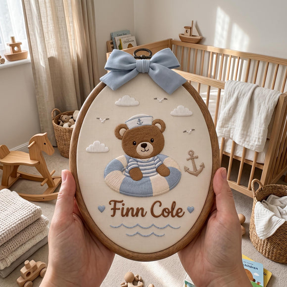 A person holds a wooden embroidery hoop featuring a personalized embroidered design of a sailor-themed teddy bear in a blue and white striped outfit with the name 'Finn Cole' and a light blue bow, set against a softly lit nursery background.