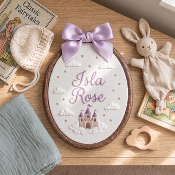 An oval wooden cross-stitch embroidery piece featuring the words 'Isla Rose' in lavender script, a lavender castle, white clouds, and small gold stars, decorated with a large lavender satin bow. The piece is styled on a wooden table with baby-themed props including a knitted bonnet, a plush bunny toy, a book, and a folded cloth.