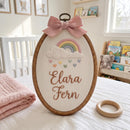 Personalized wooden embroidery hoop with a pink bow, featuring an embroidered rainbow, clouds, hearts, and the name 'Elara Fern' on a white fabric background, styled in a nursery setting.