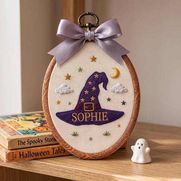 Personalized oval embroidery hanging ornament featuring a purple witch hat with gold stars, the name 'SOPHIE' in gold, embroidered clouds, stars, and a crescent moon on a cream fabric background, framed in wood with a lavender satin bow and metal ring, displayed on a wooden shelf next to books and a small white ghost figurine.