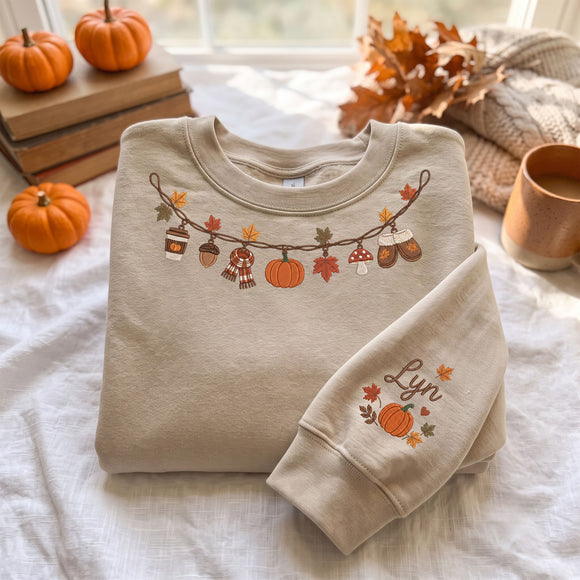 A folded beige crewneck sweatshirt with an autumn-themed graphic of pumpkins, acorns, and leaves on a string, displayed on a white bedsheet with fall-themed props including small pumpkins, books, a mug, and dried leaves.