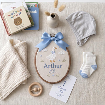 A styled baby gift set arranged on a textured cream fabric background. The central product is a personalized wooden embroidery hoop featuring a blue ribbon bow, embroidered with a duck and the name 'Arthur Eli'.