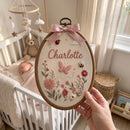 A hand holds an oval embroidered nursery wall hanging in a wooden hoop. The embroidery features the name 'Charlotte' in pink script, a pink butterfly, pink and red flowers, green leaves, ladybugs, a bumblebee, and a pink satin bow at the top.