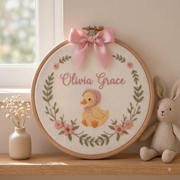 A round embroidery hoop hanging on a wooden shelf, featuring a personalized cross-stitch design with the names 'Olivia Grace' in pink, a yellow duckling in a pink bonnet, and pink floral accents. A pink satin bow is tied at the top.