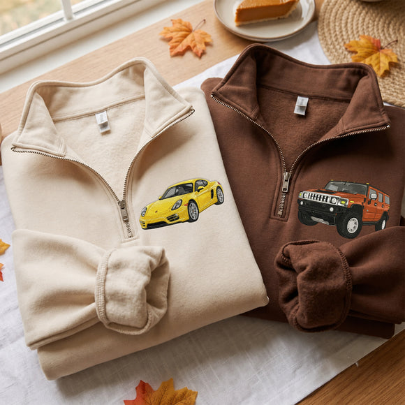 Two folded quarter-zip fleece pullovers are displayed on a table: one cream with a yellow sports car graphic and one brown with an orange SUV graphic. The scene includes autumn leaves, a slice of pumpkin pie, and a woven placemat.