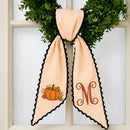 A peach and white striped autumn-themed ribbon bow with black scalloped edges, featuring a pumpkin design on one side and a monogram letter 'M' on the other, displayed on a green wreath in front of a white paneled window.