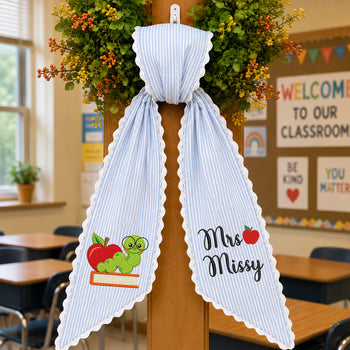 A light blue and white vertically striped bow with scalloped edges, decorated with school-themed graphics including a green caterpillar on a red apple book and the words 'Mrs. Missy' with a small red apple.