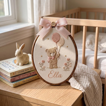 Personalized oval wooden embroidery hoop featuring a teddy bear holding pink and beige balloons with the name 'Ella' stitched below, styled on a wooden bedside table next to a crib and books.