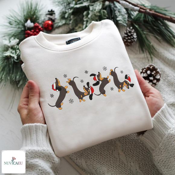 A folded cream-colored crewneck sweatshirt is held by two hands against a cozy, festive background. The sweatshirt features a graphic print of four dachshunds wearing red Santa hats and scarves, interspersed with small white snowflakes.