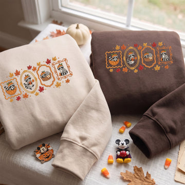 Two folded sweatshirts with autumn-themed embroidered raccoon motifs are displayed on a table near a window. One sweatshirt is cream and the other is dark brown, both featuring orange, yellow, and red fall leaves and pumpkin designs.