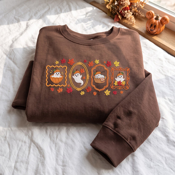 A brown crewneck sweatshirt with a Halloween-themed graphic print featuring cartoon ghosts in pumpkins and autumn leaves, laid flat on a white fabric surface with seasonal props in the background.