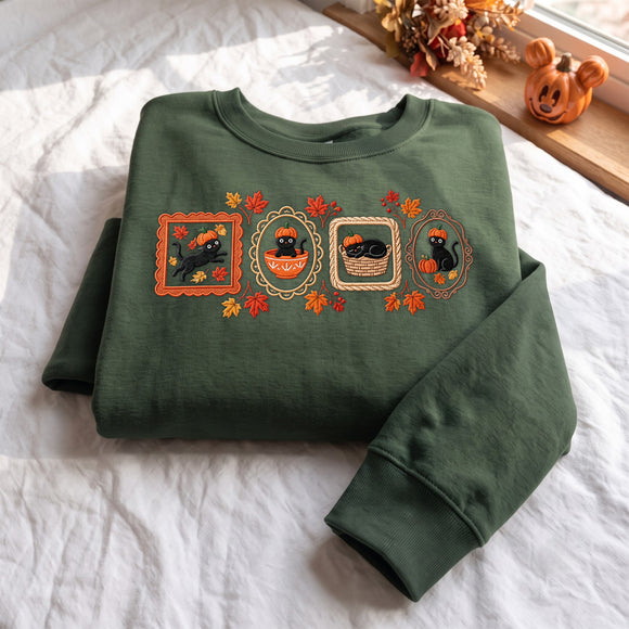 An olive green crewneck sweatshirt with a Halloween-themed graphic print featuring four black cats in autumn frames, laid flat on a white textured blanket with decorative pumpkins in the background.