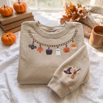 A folded beige crewneck sweatshirt with a Halloween-themed graphic print, displayed on a white fabric surface with autumn props including pumpkins, books, a mug, and a scarf.
