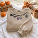 A beige crewneck sweatshirt with a graphic of a black chain holding a black cat, bat, skull, purple potion bottle, and haunted house, styled on a white fabric surface with autumn props like pumpkins, books, and a mug.