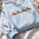 A light blue crewneck sweatshirt laid flat on a white fabric surface. The front has a colorful graphic of four dachshund-shaped Christmas vehicles, and the left sleeve has a small candy cane with holly print.