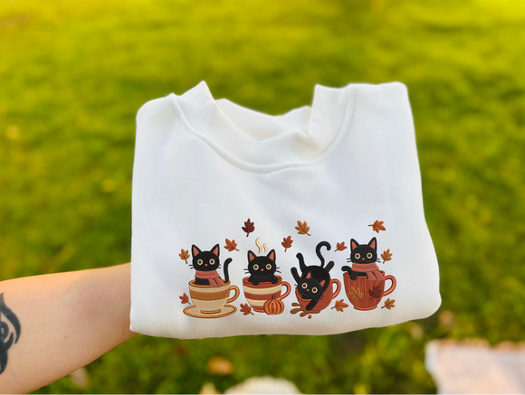 A white sweatshirt held up by a tattooed arm against a blurry green grass background. The sweatshirt features a graphic print of three black cats sitting in colorful autumn-themed cups with falling leaves.