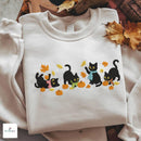 Four cute embroidered black cats wearing scarves playing with autumn leaves and pumpkins on a white sweatshirt