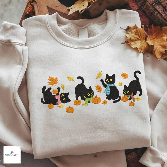 Four cute embroidered black cats wearing scarves playing with autumn leaves and pumpkins on a white sweatshirt