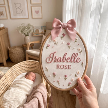 A hand holds an oval embroidered wall hanging in a wooden hoop. The embroidery features a pink satin bow at the top, small pink and white flowers, and the personalized text 'Isabelle ROSE' in pink.