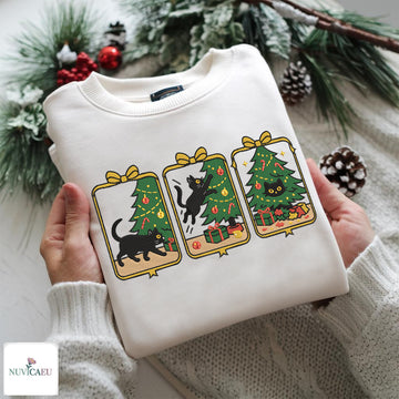 A folded white crewneck sweatshirt with a Christmas-themed graphic print of three black cats and Christmas trees, held by hands wearing a white knit sweater. The background features festive green pine branches, snow-dusted pinecones, and red berries, with a 'NUVICAEMU' brand logo in the bottom-left corner.
