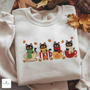 A folded off-white crewneck sweatshirt with a colorful graphic of four black cats sitting in red, yellow, and beige mugs surrounded by autumn leaves. The sweatshirt is styled on a wooden tray with scattered fall leaves, and a 'NUNICAUL' brand logo is visible in the bottom-left corner.