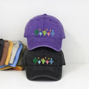 Two baseball caps displayed on a white surface against a white wall: a purple cap on top and a black cap underneath, both embroidered with colorful cactus designs. A stack of other cap colors is partially visible on the left.