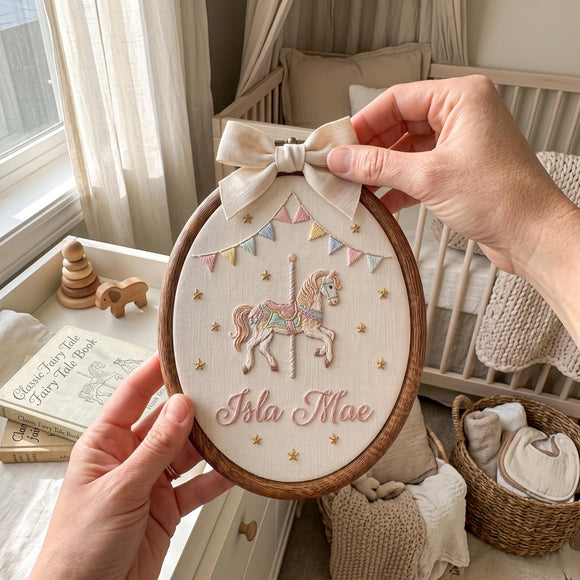 A person holds an oval embroidered nursery wall hanging in a wooden hoop. The design shows a carousel horse, pastel bunting, gold stars, and the name 'Isla Mae' in script, with a cream bow at the top.