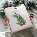 A folded cream-colored crewneck sweatshirt held by two hands, featuring a graphic of a black and white cat sitting on a green Christmas tree with colorful ornaments and presents. The background is decorated with pine branches, snow-dusted pinecones, and red berries, and a white knit sweater is draped underneath.