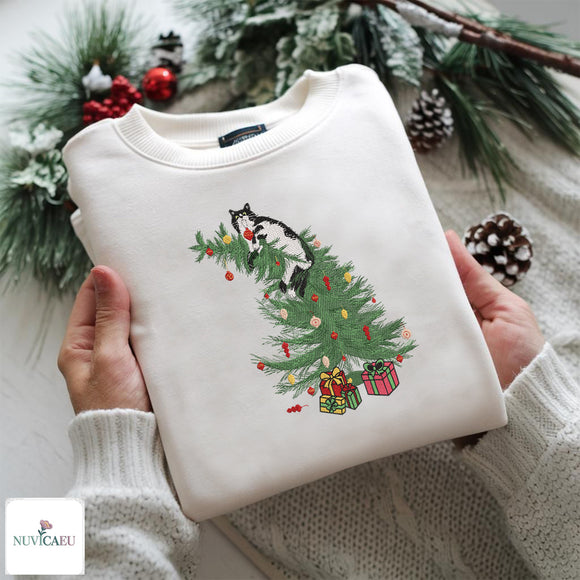 A folded cream-colored crewneck sweatshirt held by two hands, featuring a graphic of a black and white cat sitting on a green Christmas tree with colorful ornaments and presents. The background is decorated with pine branches, snow-dusted pinecones, and red berries, and a white knit sweater is draped underneath.