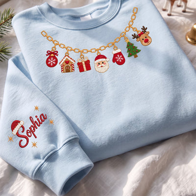 A light blue crewneck sweatshirt laid flat on a white fabric surface. The sweatshirt features a gold chain graphic across the chest with embroidered Christmas-themed charms including a gingerbread house, gift box, Santa face, Christmas tree, and reindeer.