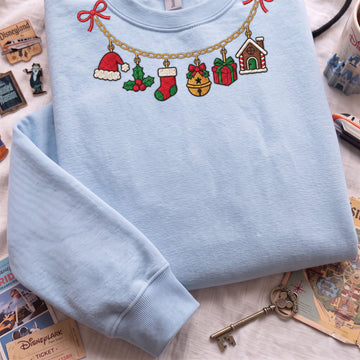 A light blue crewneck sweatshirt laid flat on a white surface, decorated with colorful embroidered Christmas-themed motifs along a gold chain, including a Santa hat, stocking, bell, gift, and gingerbread house. Disney-themed props and a vintage key surround the sweatshirt.